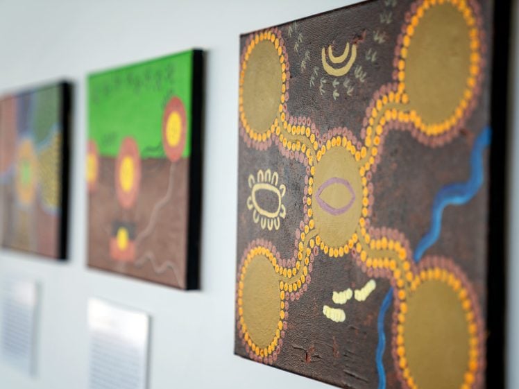 Image of Close-up of colourful Indigenous Australian dot paintings on a wall, featuring circular patterns, wavy lines, and traditional symbols in earthy tones with a green and brown background.