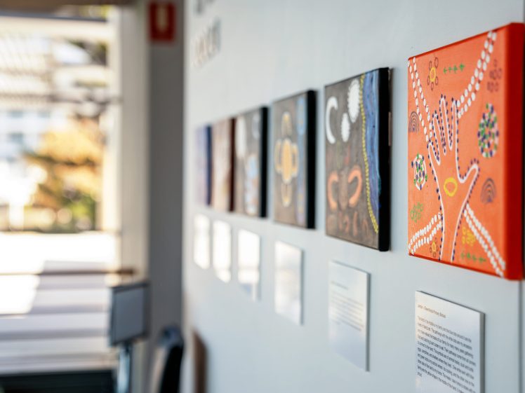 Image of A row of small, colourful artworks is displayed on a white gallery wall, with the nearest piece featuring a hand design on an orange background; descriptive labels are mounted below each artwork.