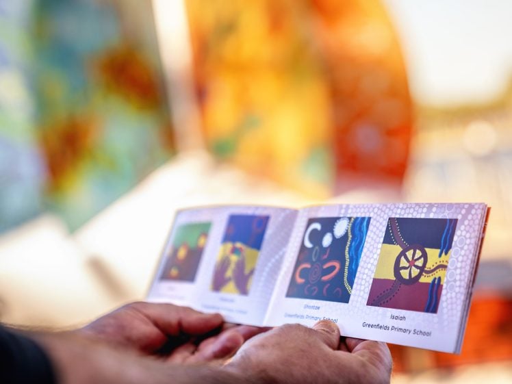Image of A person holds an open booklet displaying colourful children's artwork, with blurred, vibrant art installations in the background, outdoors in bright sunlight.