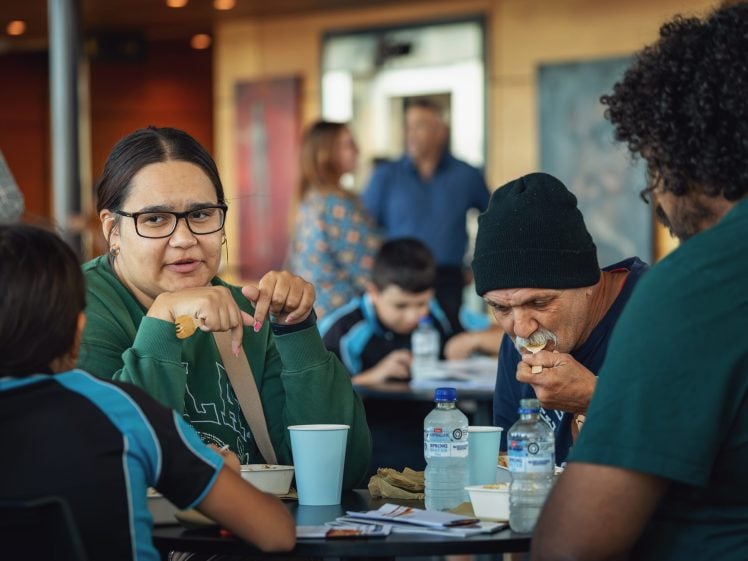 Image of A woman in glasses and a green jumper sits at a table, talking while others eat. Two children and two men are also at the table, with water bottles, cups, and papers scattered around. The background shows more people and colourful walls.