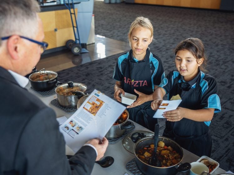 Image of Two children in uniforms serve food from pots on a table to an adult holding papers with recipes or instructions. The scene appears to be an event or cookery activity in a spacious indoor setting.