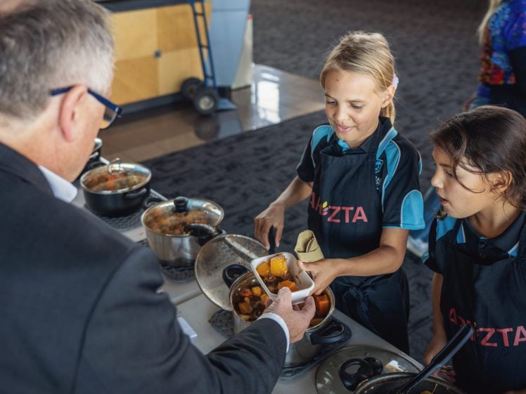 Image of Two young girls in aprons serve food from pots to a man in a suit across a table. One girl holds a bowl whilst the other ladles stew into it. The scene appears to be a food event or cookery competition.