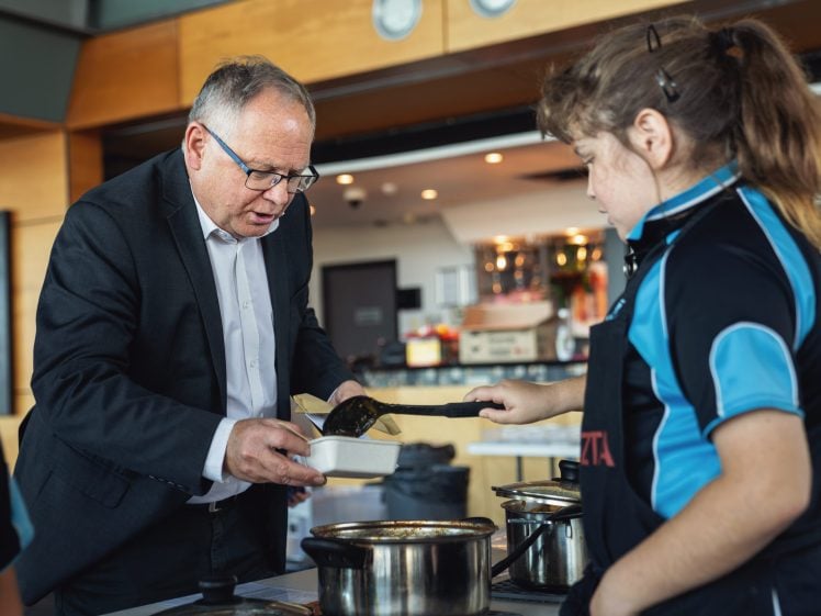 Image of An older man in a suit helps a young girl in a school uniform cook at a cooker, stirring food in a saucepan while another child looks on. They appear to be in a kitchen or cookery classroom.