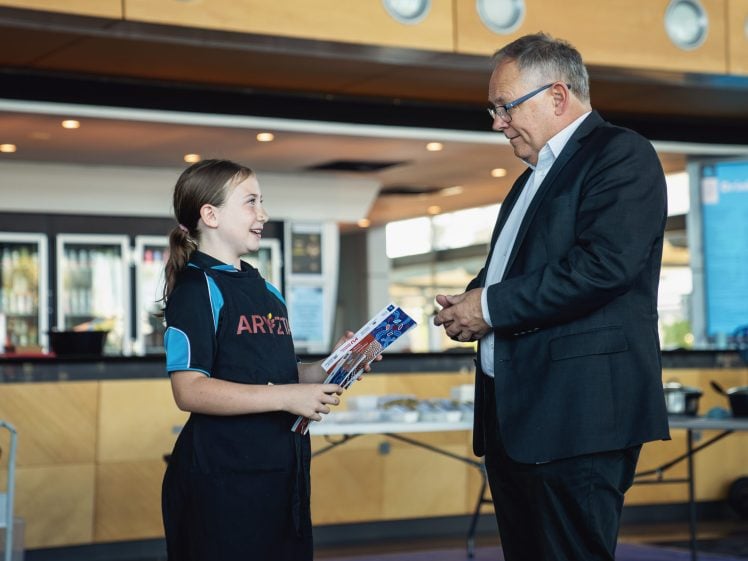 Image of A young girl in a dark pinafore smiles whilst holding kitchen utensils and speaking with an older man in a suit inside a modern, well-lit room with a counter and shelves in the background.