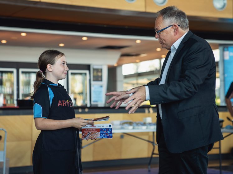 Image of A young girl in a sports kit stands holding a booklet while an older man in a suit gestures towards her, both engaged in conversation inside a brightly lit room.