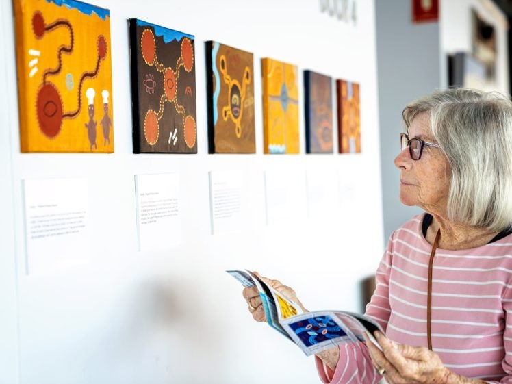 Image of An older woman with grey hair and glasses, wearing a pink striped shirt, holds a brochure while viewing colourful abstract paintings displayed on a white gallery wall.