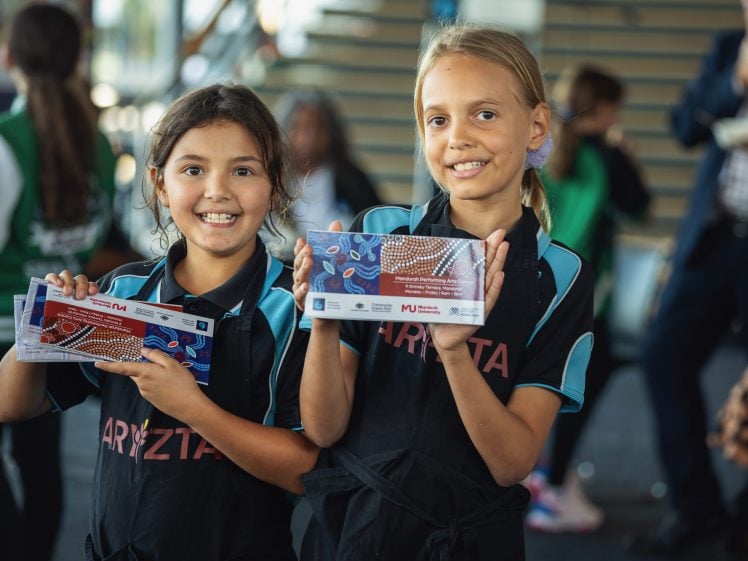 Image of Two smiling girls in matching uniforms hold up colourful rectangular items with patterns, possibly prizes or certificates, standing indoors among other people.