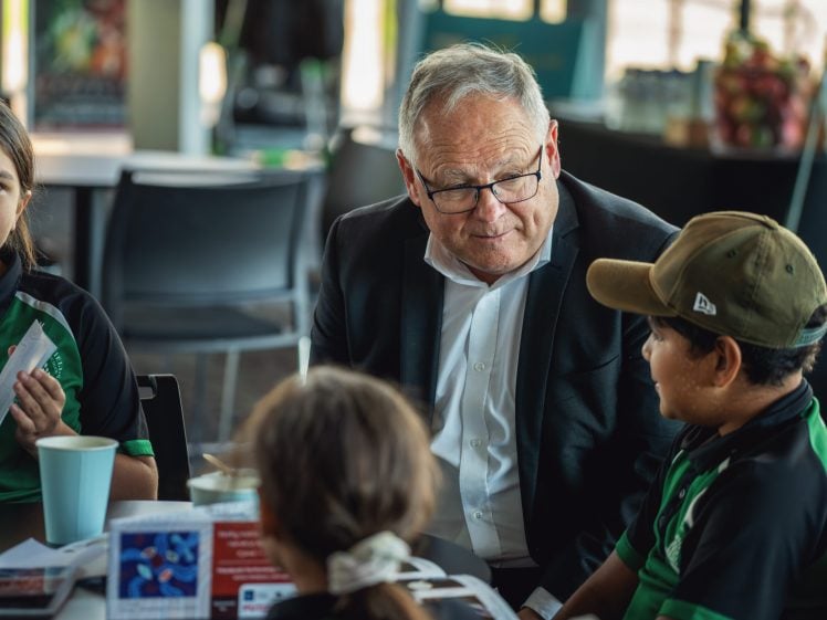 Image of An older man in a suit and glasses sits at a table with three children, engaging in conversation. The group appears to be indoors, and the children are wearing green and black uniforms. Papers and cups are on the table.