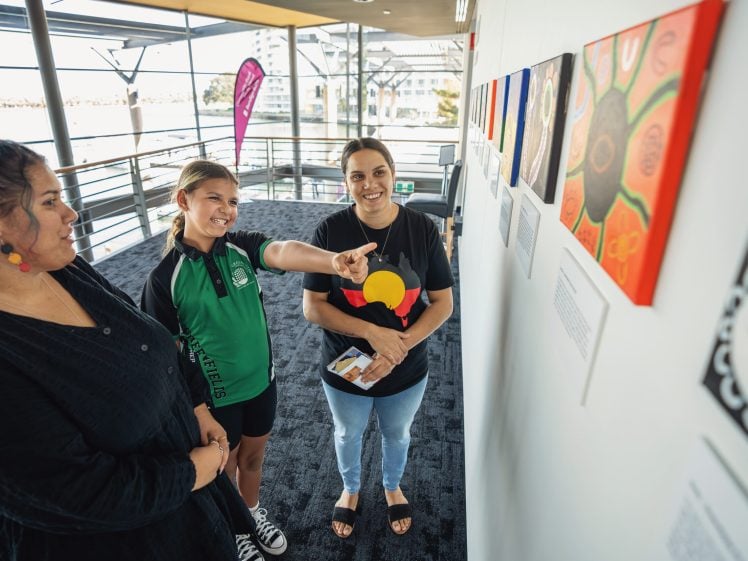 Image of Three people stand in a bright corridor, looking at colourful artwork displayed on a wall. One person points at a painting while the others smile, engaging in discussion. Large windows provide natural light in the background.