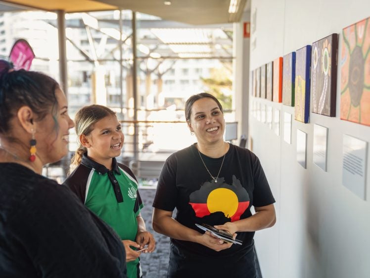 Image of Three people stand indoors, smiling and looking at colourful artwork displayed on a wall. One person wears a shirt featuring the Aboriginal flag. The setting is bright with large windows in the background.