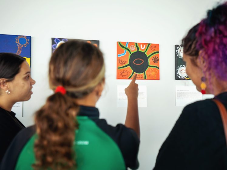 Image of Three people stand in front of colourful artwork displayed on a wall. One person in the centre, wearing a green top, points at an orange and black painting while the others look on attentively.