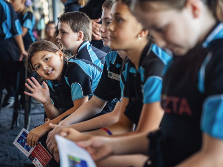 Image of A group of children in matching blue and black uniforms sit in a row. One girl in the foreground smiles and waves at the camera, whilst others look ahead or hold papers. The atmosphere appears lively and friendly.