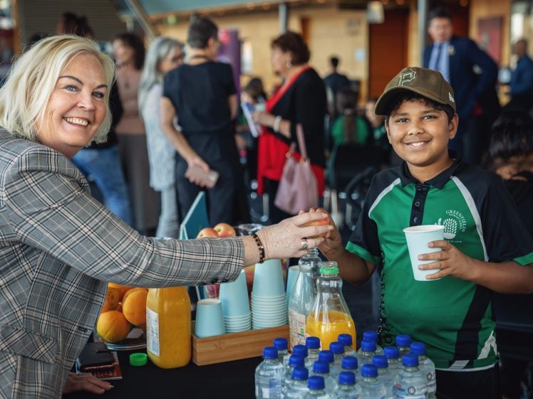 Image of A smiling woman in a checked blazer and a smiling boy in a green polo shirt and cap shake hands at a refreshments table with juice, water, and fruit; people chat in the background.