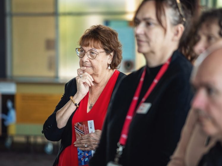 Image of A group of adults stands indoors, with a woman in a red top and glasses thoughtfully resting her hand on her chin in the foreground. The background is softly focussed.