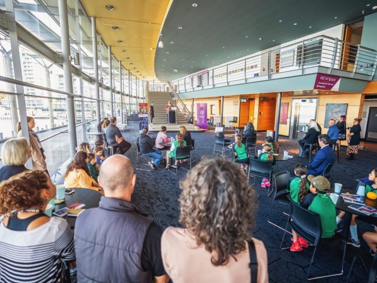 Image of A group of people sit and stand in a bright, modern indoor space with large windows and a curved ceiling, watching a presentation or event near a staircase. Some children wear green shirts and hats.