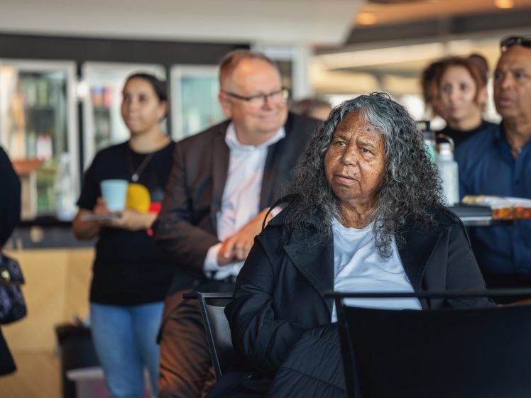 Image of A woman with long, curly grey hair sits in the foreground at an indoor event, looking ahead. Several people are seated and standing in the background, some holding drinks and food, in a casual, modern setting.