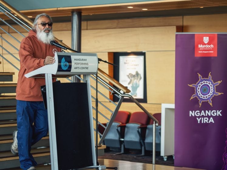 Image of An older man with a grey beard, sunglasses, and an orange hoodie speaks at a podium in the Mandurah Performing Arts Centre. A purple Ngangk Yira banner stands nearby. Modern interior with staircase and seating in the background.