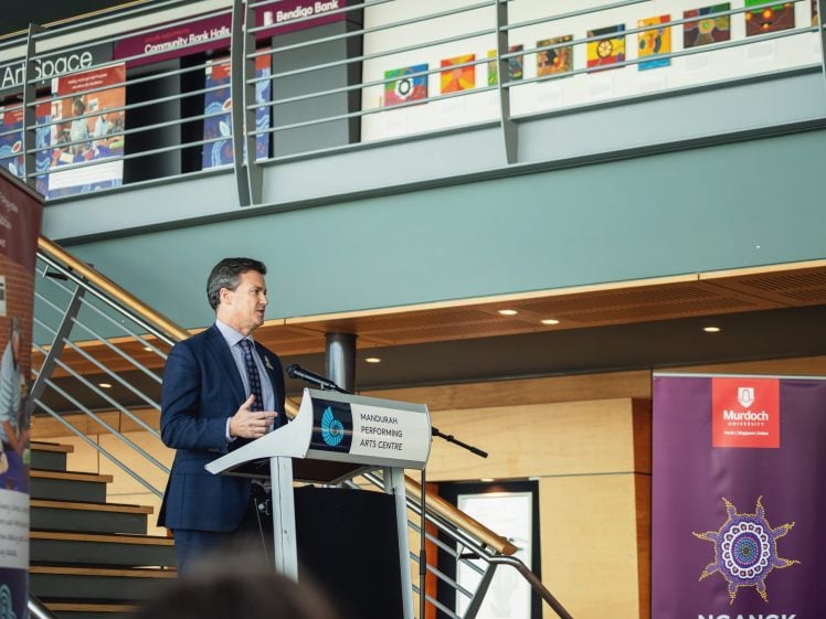 Image of A man in a suit speaks at a lectern inside a modern building, with banners and artwork displayed in the background and people seated in the foreground.
