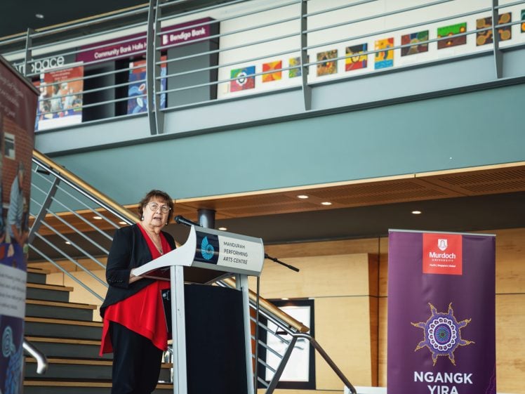 Image of A woman in a red top and black jacket speaks at a lectern in a modern building, with stairs and colourful artwork displayed on the upper level. Banners for Ngangk Yira and Murdoch University are visible beside her.