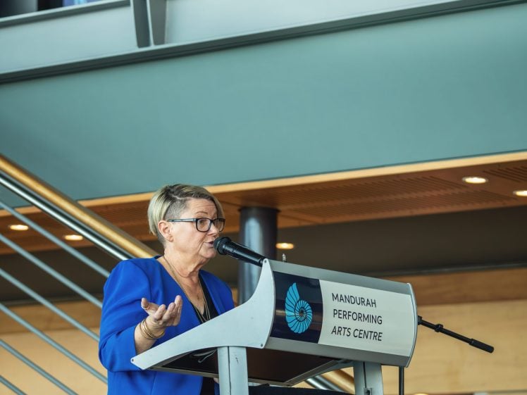 Image of A woman wearing glasses and a blue blazer speaks at a lectern labelled Mandurah Performing Arts Centre in an indoor venue with stairs and artwork on the wall behind her.