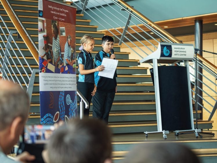 Image of Two people in uniforms stand on stairs, speaking into a microphone and holding papers at a lectern labelled Mandurah Performing Arts Centre, whilst an audience looks on. A colourful banner is displayed beside them.