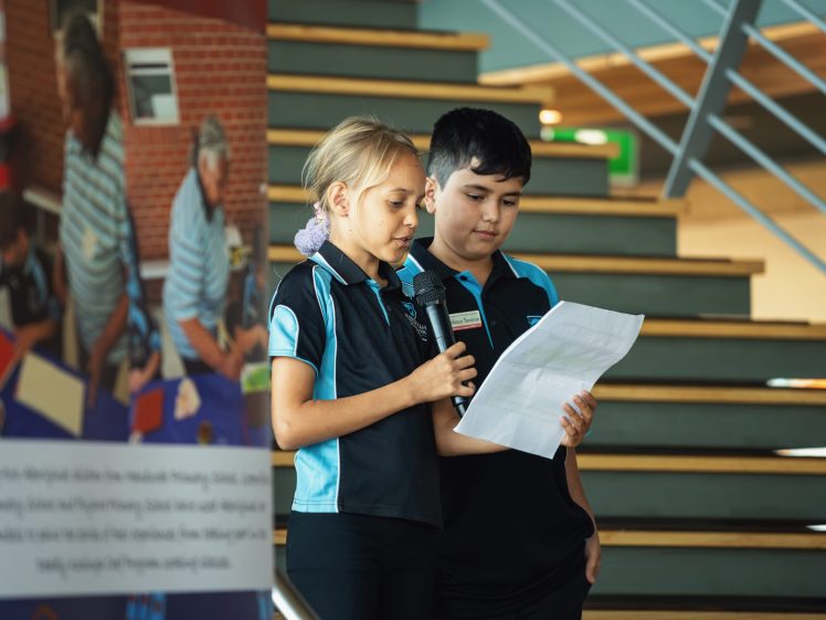 Image of Two children in school uniforms stand on stairs; one holds a microphone and reads from a piece of paper while the other looks on. A blurred banner and an adult reading to children are visible in the background.