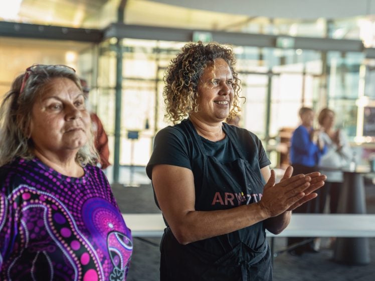 Image of Two women, one in a colourful shirt and one in a black apron, stand indoors. The woman in the apron is clapping and smiling, whilst others in the background watch and applaud in a bright, modern space.