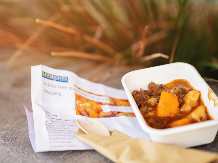 Image of A disposable container filled with stew containing meat, potatoes, and carrots sits next to a printed recipe and a wrapped wooden fork and knife, outdoors on a concrete surface.
