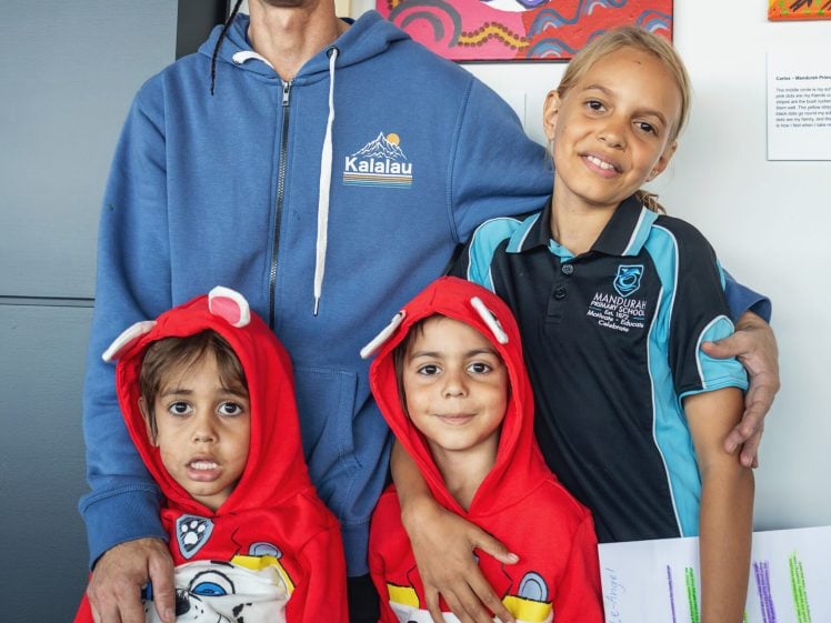 Image of A man stands smiling with three children in front of colourful artwork. Two young children wear matching red hoodies; an older girl in a school uniform stands beside them, holding papers and smiling.