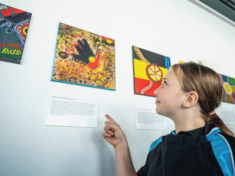 Image of A young girl points at a colourful painting displayed on a white gallery wall, with other vibrant artworks visible beside it and description cards mounted below the paintings.