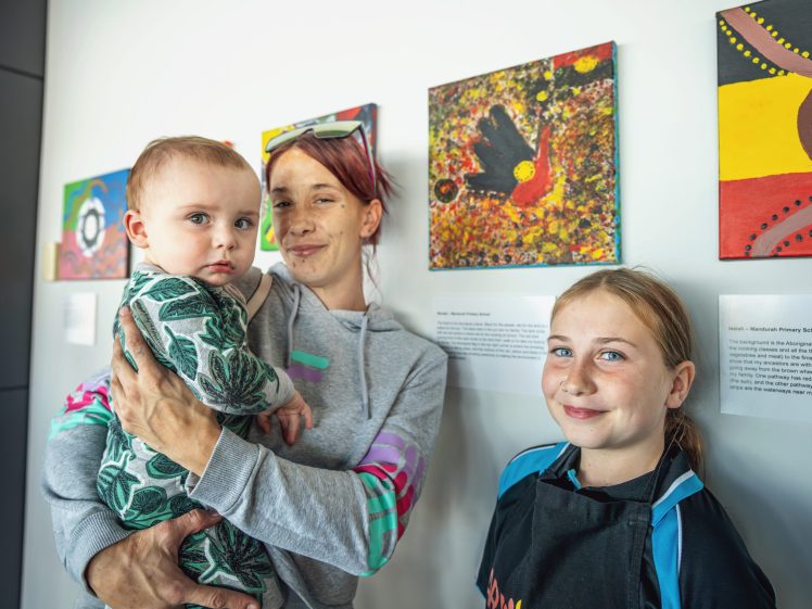 Image of A woman holds a baby while standing next to a smiling girl in front of colourful artwork on display in a gallery. The wall behind them features paintings and descriptive labels.