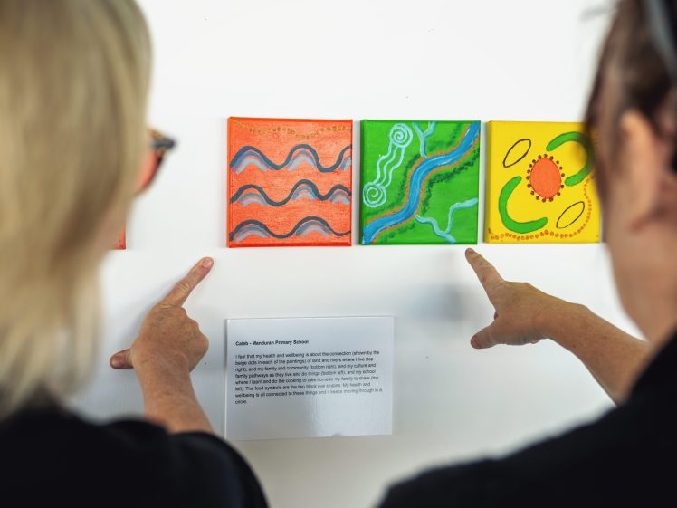 Image of Two people stand in front of a wall, each pointing at colourful, abstract paintings. A small plaque with descriptive text is mounted below the artwork. Their faces are out of focus and turned away from the camera.