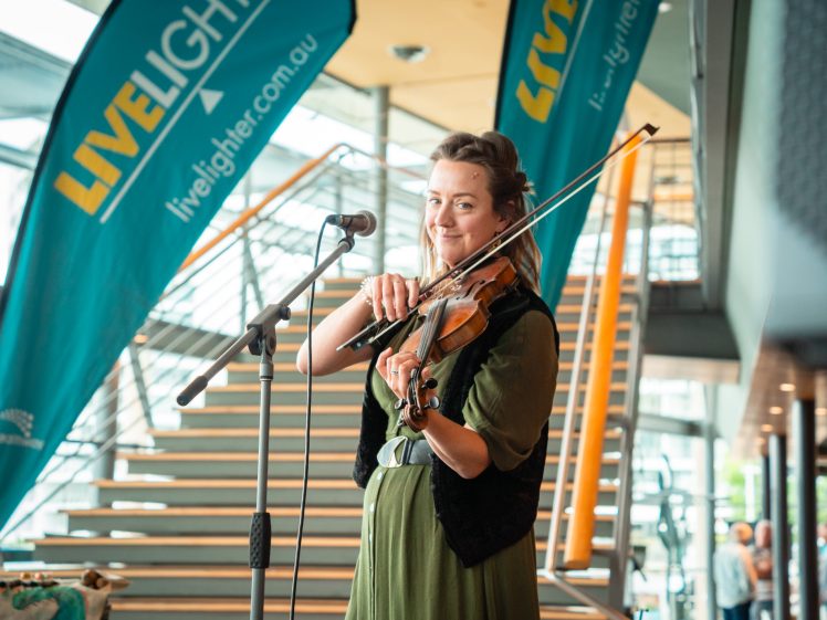 Image of A woman in a green dress plays the violin and smiles at the camera, standing near a microphone. Behind her are stairs and large blue LiveLighter banners at an indoor event.