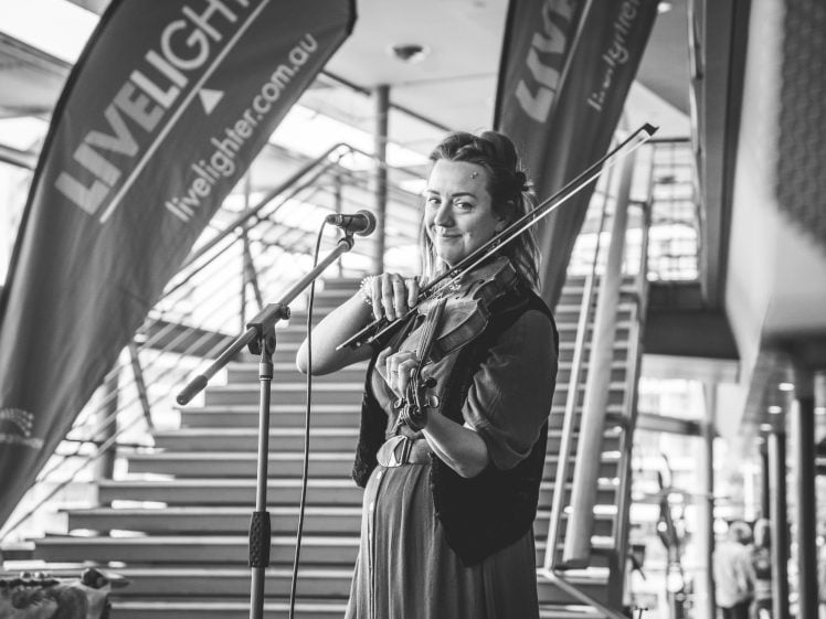 Image of A woman stands in front of a microphone playing a violin and smiling. Behind her, there are large LiveLighter banners and a staircase in a modern indoor setting. The image is in black and white.