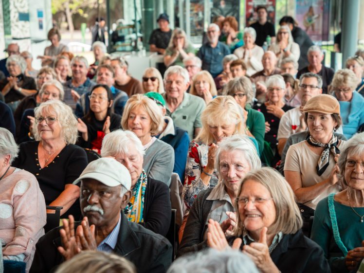 Image of A large group of older adults, both men and women, sit closely together indoors, smiling and applauding as they attend an event or presentation. The atmosphere appears cheerful and engaged.
