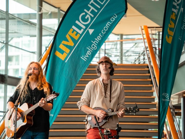 Image of Two musicians perform indoors in front of a staircase; one plays an acoustic guitar and sings, the other plays a mandolin. Teal “LiveLighter” banners are displayed behind them.