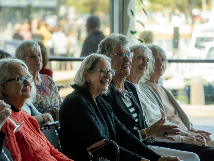 Image of A group of older women sit closely together indoors, smiling and enjoying an event. Sunlight streams through large windows behind them, revealing a marina with boats outside.