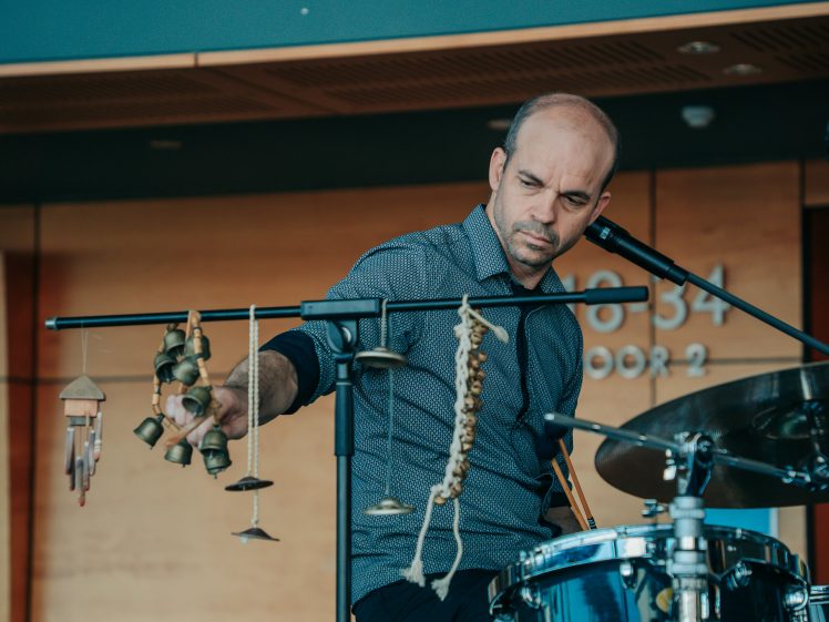 Image of A man sits at a drum kit, holding small percussion bells and chimes attached to a horizontal bar. He appears focused as he performs on stage, with a microphone and wooden backdrop visible behind him.
