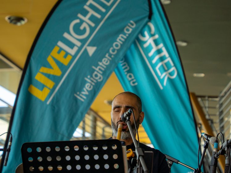 Image of A musician sings into a microphone behind a music stand, with large blue banners reading LIVELIGHTER livelighter.com.au displayed in the background.