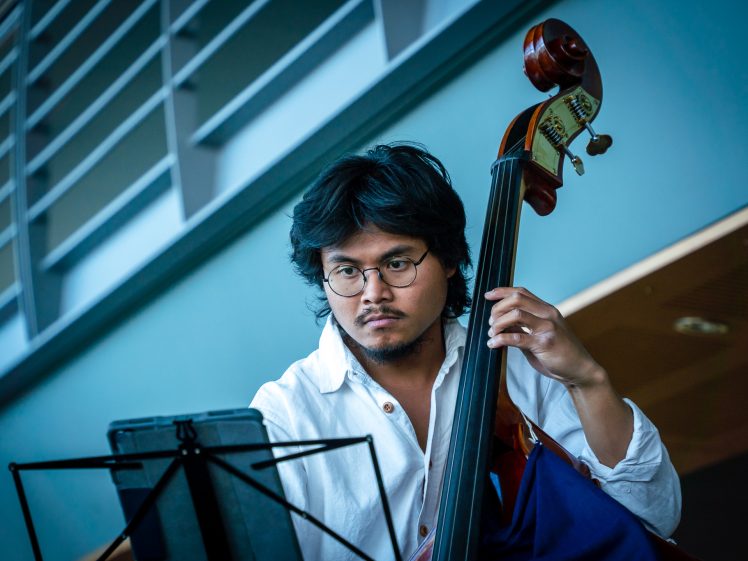 Image of A man with glasses plays a double bass while reading sheet music. He is wearing a white shirt and appears focused, with a modern interior background behind him.