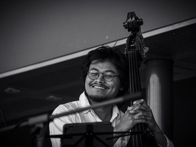 Image of A musician with glasses and a beard smiles warmly whilst holding a double bass, performing indoors. The image is black and white, capturing a joyful moment during the performance.