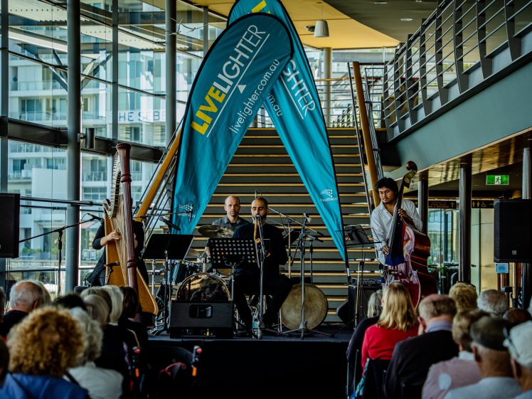 Image of A jazz band performs on a small indoor stage in front of an audience, with two large blue LiveLighter banners and a staircase in the background. The band includes a harpist, drummer, and double bassist.