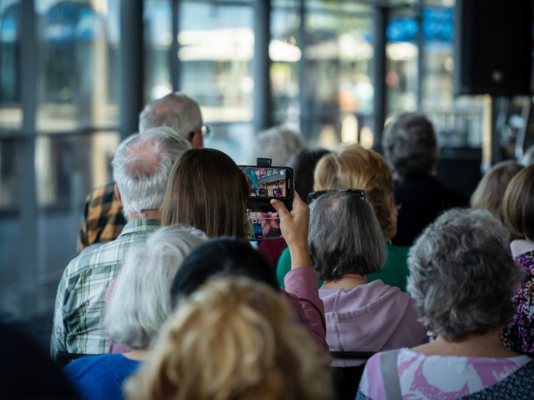 Image of A group of older adults sits closely together indoors, facing forward. One person in the middle raises a mobile phone, capturing a photo or video of the event. The image is bright, with large windows in the background.