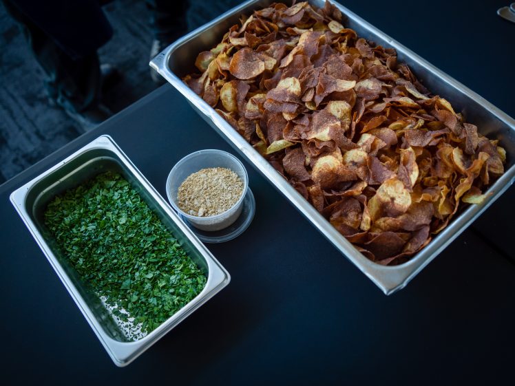 Image of A metal tray of crispy potato crisps sits next to containers of chopped fresh herbs and grated cheese on a dark table.