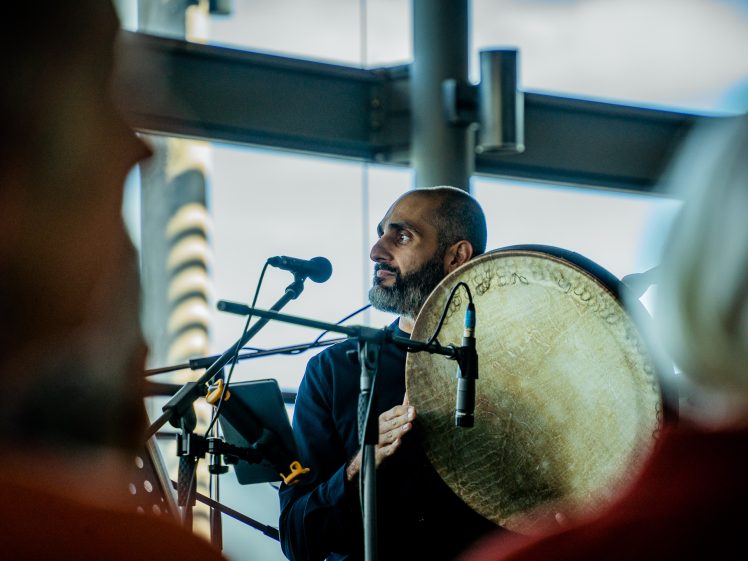 Image of A man plays a large frame drum and sings into a microphone at an indoor event, with blurred audience members in the foreground and natural light coming through tall windows behind him.