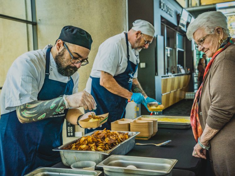 Image of Two chefs in blue aprons serve food from trays to an elderly woman at a counter in a brightly lit indoor setting. One chef is serving food, while the woman waits with a bowl in hand.