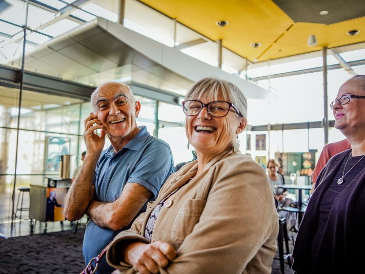 Image of Three older adults stand indoors, smiling and talking together. The man on the left has his hand near his face, whilst the two women beside him are laughing. The setting is bright with large windows in the background.