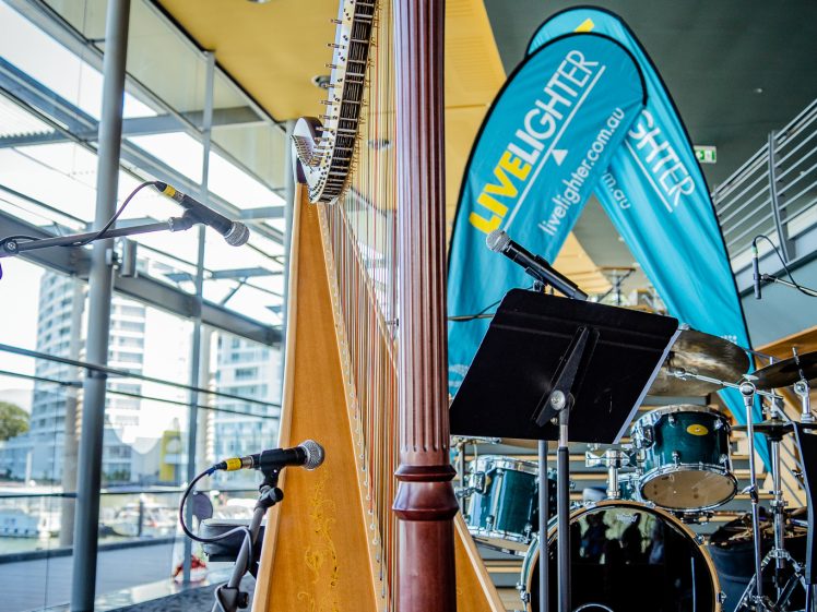 Image of A wooden harp stands on a stage near microphones, drums, and blue Live Lighter banners, with large windows showing buildings outside.