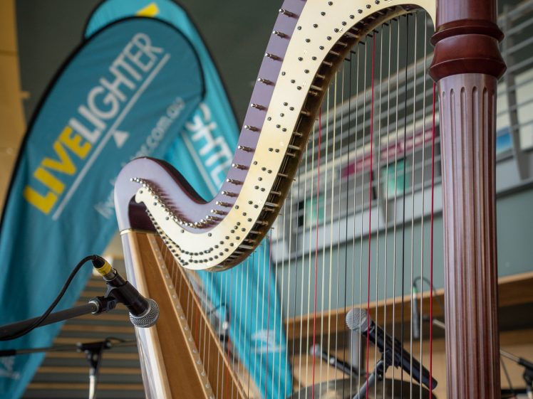 Image of A close-up of a wooden harp with strings, set on a stage. Behind the harp are a microphone, drums, and a blue banner with the text LIVE LIGHTER. Steps and a railing are visible in the background.