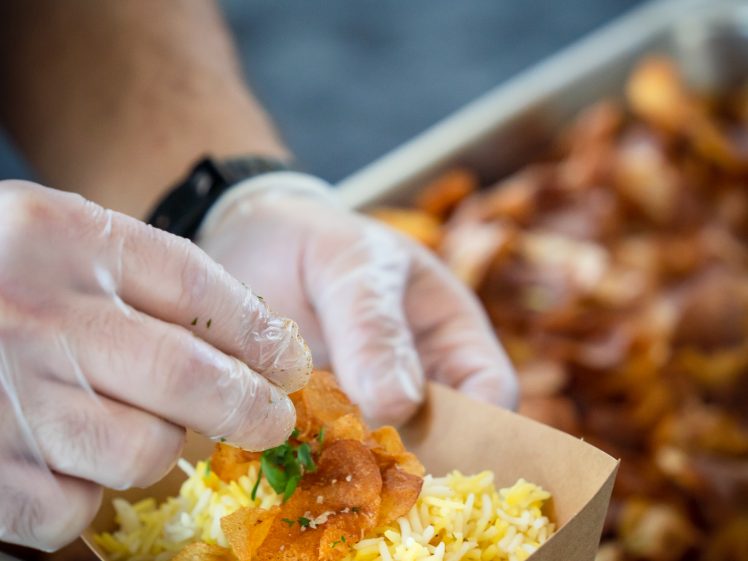 Image of A person wearing gloves garnishes a serving of yellow rice with crispy crisps and herbs in a brown paper tray, with more crisps visible in a large metal container nearby.